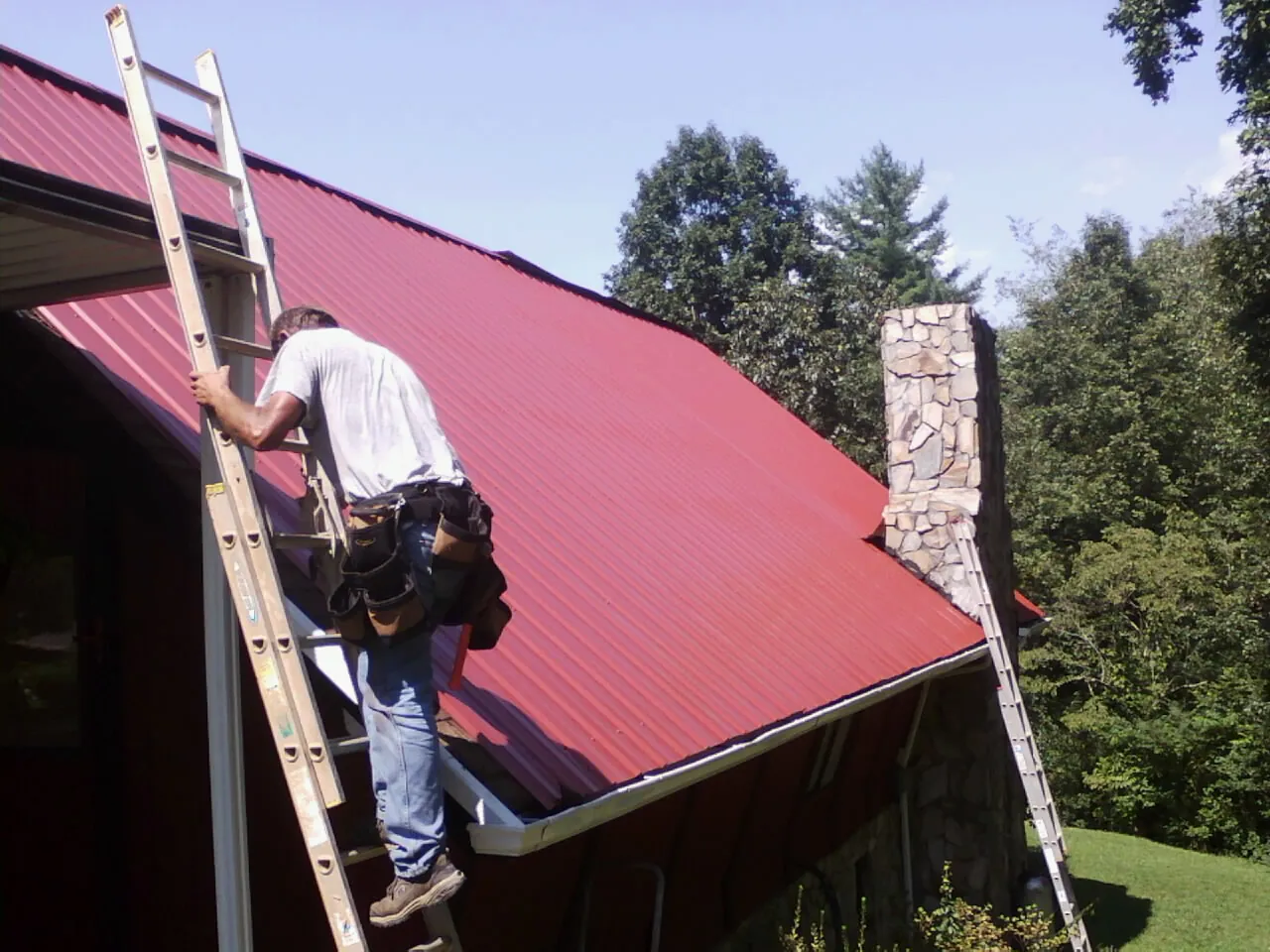 Roofer on a ladder beside a red metal roof and stone chimney — Soffit & Fascia Repair by Roofing Brothers in Wild Rose